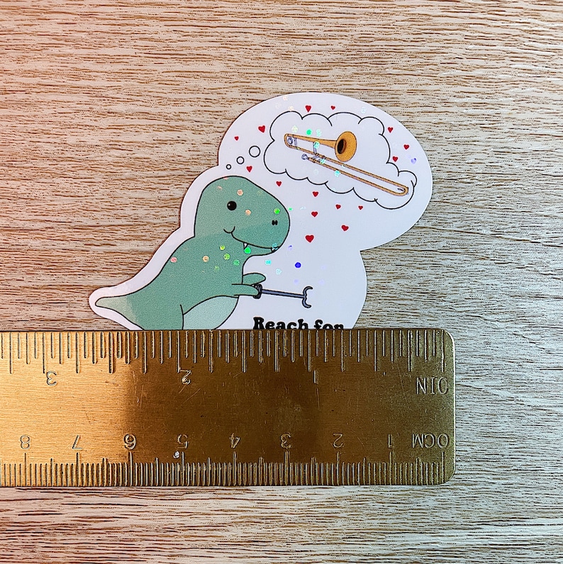 May include: A green dinosaur sticker with glitter accents holding a trombone. The dinosaur is looking up at a thought bubble with a gold trombone inside. The thought bubble has red hearts around it. The sticker says "Reach for" at the bottom.