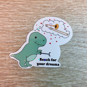 May include: A green dinosaur holding a trombone with a thought bubble above its head showing a gold trombone. The text below the dinosaur reads "Reach for your dreams".