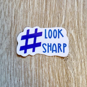 May include: A die-cut sticker with a white background featuring a large blue hashtag symbol on the left side. To the right of the symbol, the text "LOOK SHARP" is printed in blue capital letters. The sticker has an irregular, rounded edge shape.