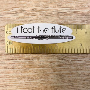 May include: White oval sticker with the text "i toot the flute" and a black and white illustration of a flute. The sticker is placed on a gold ruler.