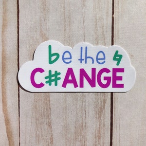 May include: A white sticker in the shape of a cloud with the words "be the C#ANGE" in green, blue, and purple. The sticker is on a light brown wooden surface.
