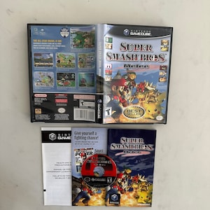 Könnte beinhalten: Ein Nintendo GameCube-Spielecase und eine Disc für Super Smash Bros. Melee. Das Case ist schwarz mit einem roten und weißen Etikett, auf dem der Titel des Spiels und die Charaktere abgebildet sind. Die Disc ist schwarz mit einem roten Etikett, auf dem der Titel des Spiels und die Charaktere abgebildet sind. Das Case enthält außerdem ein kleines Heft mit Gesundheits- und Sicherheitshinweisen.