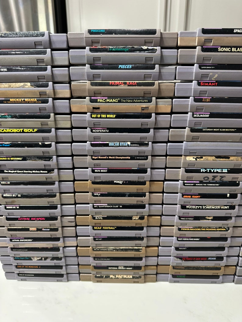 May include: A stack of over 30 vintage Super Nintendo Entertainment System (SNES) game cartridges. The cartridges are arranged in rows and columns, with the titles facing forward. Some of the titles include Mickey Mania, Pac-Man 2, Primal Rage, and R-Type III.