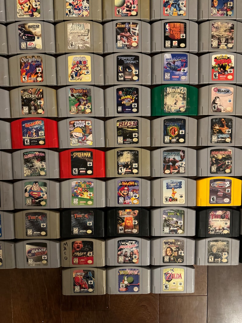 May include: A collection of 64 Nintendo 64 game cartridges, arranged in a grid pattern. The cartridges are in various colors, including gray, black, red, green, and yellow. The cartridges are arranged in rows of eight, with each row containing a different game title. Some of the game titles include "StarCraft", "Spiderman", "Smash Bros", "Zelda", "Quake II", "Rush", "Perfect Dark", and "Paper Mario".