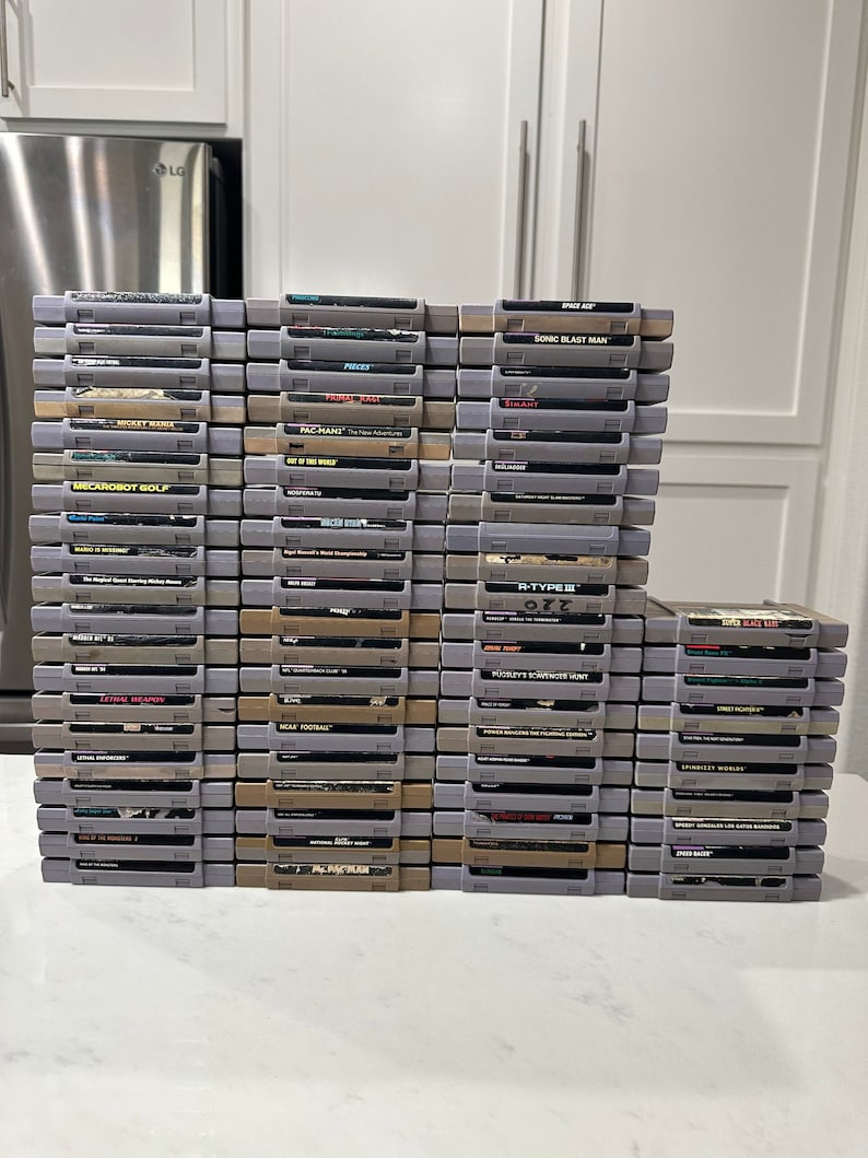 May include: A stack of over 40 vintage Super Nintendo Entertainment System (SNES) game cartridges. The cartridges are arranged in rows and columns, with the game titles visible on the front of each cartridge. Some of the titles include "Sonic Blast Man", "R-Type III", "Pac-Man", "NCAA Football", and "SimAnt".