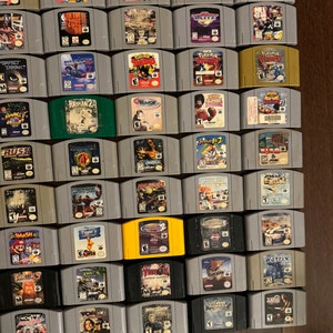 May include: A collection of 48 Nintendo 64 game cartridges, including titles like "StarCraft", "Smash Bros", "Rayman 2", "Zelda", "WCW/nWo Revenge", and "South Park", displayed in rows on a wooden surface.