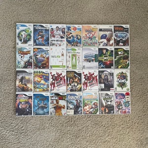 May include: A collection of 24 video game cartridges for the Nintendo Wii console. The cartridges are arranged in four rows of six. The games include titles such as Rayman, FIFA Soccer 11, Wii Fit, and High School Musical.