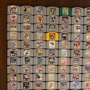 May include: A collection of 64 Nintendo 64 game cartridges, arranged in a grid pattern on a wooden surface. The cartridges are mostly gray with colorful labels featuring various game titles, including "GoldenEye 007", "Super Mario 64", "The Legend of Zelda: Ocarina of Time", and "Star Fox 64".