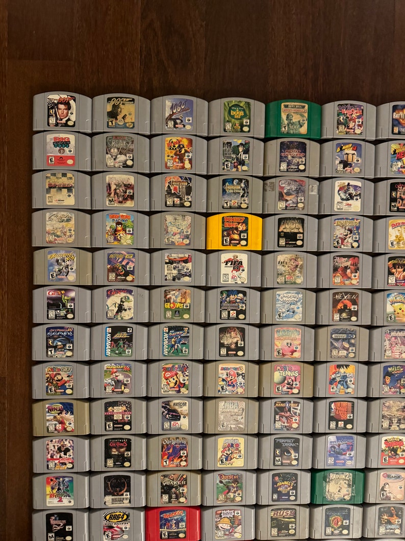 May include: A collection of 64 Nintendo 64 game cartridges, arranged in a grid pattern on a wooden surface. The cartridges are mostly gray with colorful labels featuring various game titles, including "GoldenEye 007", "Super Mario 64", "The Legend of Zelda: Ocarina of Time", and "Star Fox 64".