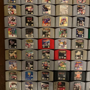 May include: A collection of 48 Nintendo 64 game cartridges, arranged in a grid on a wooden surface. The cartridges are in various colors, including gray, black, red, and blue. Some of the cartridges have titles visible, such as "Mario Kart 64", "Ocarina of Time", and "GoldenEye 007".