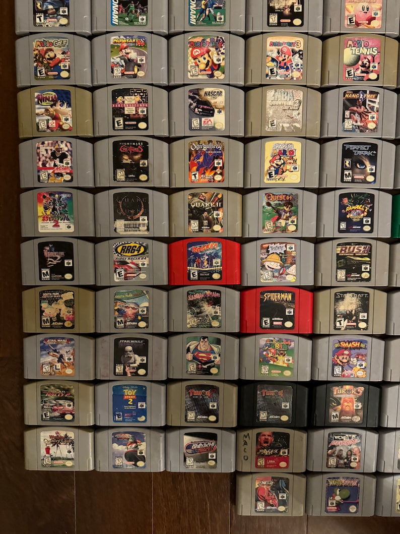 May include: A collection of 48 Nintendo 64 game cartridges, arranged in a grid on a wooden surface. The cartridges are in various colors, including gray, black, red, and blue. Some of the cartridges have titles visible, such as "Mario Kart 64", "Ocarina of Time", and "GoldenEye 007".