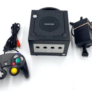 Puede incluir: Una consola Nintendo GameCube negra con un mando a juego, un adaptador de corriente y cables AV. La consola tiene el logotipo de Nintendo GameCube en la parte superior. El mando tiene una variedad de botones de colores.