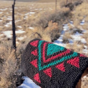 May include: A black saddle pad with a geometric design in red and teal. The pad is resting on a wooden saddle rack outdoors, with a backdrop of a field and mountains.