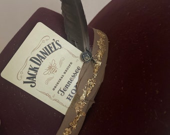 Pode incluir: Um chapéu de feltro bordô com uma fita de guarnição dourada e marrom. O chapéu tem uma pena no topo e um rótulo que diz "Jack Daniel's Original Recipe Tennessee Honey".