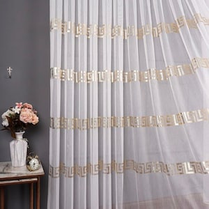 Puede incluir: Cortinas blancas de voile con ribete dorado con patrón de llave griega. Las cortinas están colgadas en una habitación con una pared gris y una mesa auxiliar con un jarrón de flores.