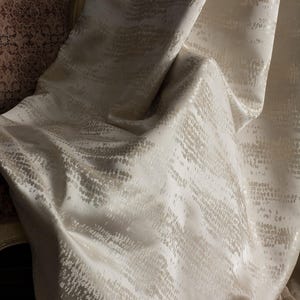 May include: Off-white fabric with a subtle, textured pattern. The fabric drapes in soft folds, catching the light and creating highlights and shadows. The pattern appears to be a series of small, irregular dashes, adding visual interest to the textile.