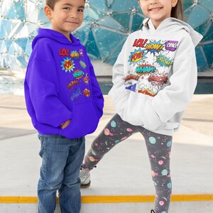 Könnte beinhalten: Zwei Kinder tragen Hoodies mit bunten Comic-Soundeffekten, die darauf gedruckt sind. Der Junge trägt einen lila Hoodie und eine blaue Jeans. Das Mädchen trägt einen weißen Hoodie und eine graue Leggings mit einem Weltraumdruck.