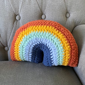 Könnte beinhalten: Ein regenbogenfarbenes Häkelkissen in Form eines Bogens. Das Kissen ist aus weichem, flauschigem Garn gefertigt und zeigt Farbtöne von Rot, Orange, Gelb, Grün, Blau und Lila.