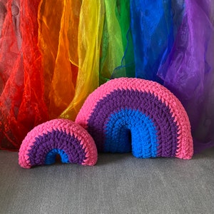 May include: Two crocheted rainbow-shaped pillows, one larger than the other, with pink, purple, and blue yarn. The pillows are sitting on a gray surface.