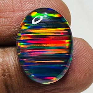 Könnte beinhalten: Ein ovaler, polierter Edelstein mit einer lebendigen Farbpalette. Der Stein hat eine dunkle Basis mit horizontalen Bändern in Rot, Gelb, Grün und Blau, die einen Regenbogeneffekt erzeugen. Der Edelstein wird zwischen Fingern gehalten.