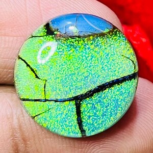 May include: A round, dichroic glass cabochon with a vibrant green and blue color scheme. The surface has a textured appearance with black crackle lines. The top edge features a blue section, and the overall design is reminiscent of a landscape.