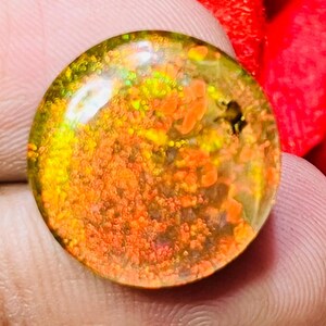 May include: A round, polished gemstone with a vibrant display of colors. The gem features a mix of orange, yellow, and green hues, with a speckled pattern. The gemstone is held against a red background.
