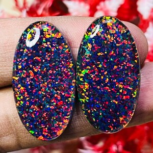 Puede incluir: Dos piedras preciosas ovaladas con una base azul oscuro y una vibrante exhibición de motas coloridas. Las piedras preciosas se sostienen en una mano, mostrando su superficie pulida y el juego de la luz sobre el diseño moteado. Las piedras son probablemente ópalos.