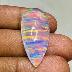Könnte beinhalten: Ein tropfenförmiger Opal-Cabochon mit einer weißen Basis und schillernden Regenbogenfarben. Der Opal hat ein einzigartiges Muster aus Streifen und Wirbeln.