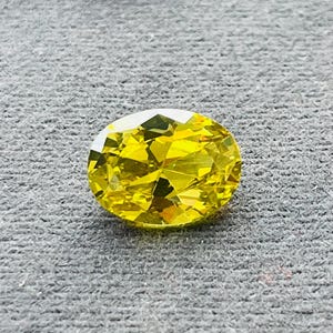 Puede incluir: Una piedra preciosa amarilla facetada de forma ovalada. La gema tiene un corte brillante, que refleja la luz con múltiples facetas. La piedra se coloca sobre un fondo gris, resaltando su color vibrante y claridad. Esta piedra preciosa es adecuada para joyería.