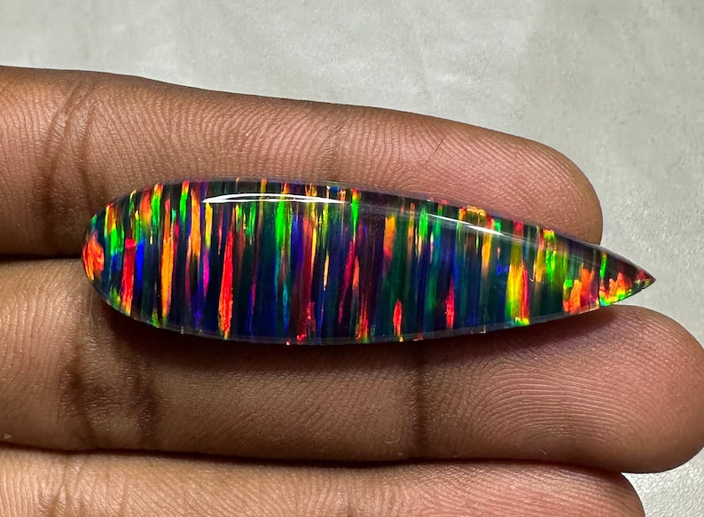 May include: A black opal cabochon with a teardrop shape and a vibrant display of iridescent colors, including red, orange, yellow, green, blue, and purple.