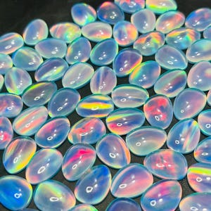 Peut inclure: Une collection de pierres précieuses ovales et irisées. Les pierres présentent une gamme de couleurs, notamment des bleus, des roses et des jaunes, avec un effet chatoyant et opalescent. Les pierres sont dispersées sur une surface sombre.