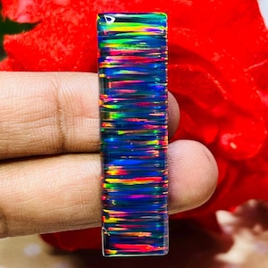 Puede incluir: Una piedra preciosa rectangular y pulida con una vibrante exhibición de color. La piedra presenta una base azul oscuro con bandas horizontales rojas, amarillas y verdes, creando un efecto iridiscente. Es probable que sea un ópalo.