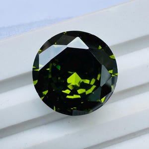 May include: A round, faceted gemstone with a deep forest green color and bright lime green highlights. The stone features a complex geometric cut with multiple triangular and trapezoidal facets that reflect light, creating a high-contrast appearance.