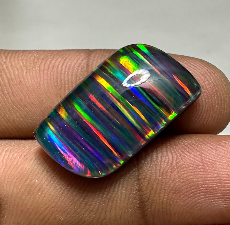 Op de afbeelding: Een donkere, iriserende opaal cabochon met een gestreept patroon van groen, blauw, rood en geel. De opaal heeft een glad, gepolijst oppervlak.