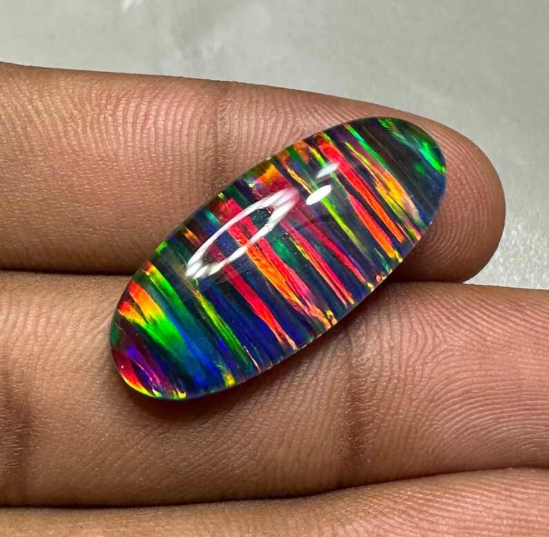 May include: A colorful, iridescent opal cabochon with a striped pattern in shades of red, orange, green, blue, and black. The opal is oval-shaped and has a smooth, polished surface.