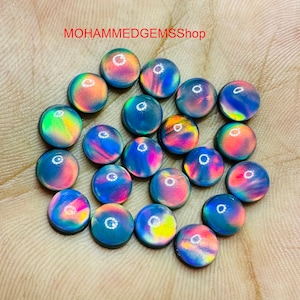 May include: A collection of 20 round, iridescent opal cabochons. The opals are a mix of blue, green, pink, and yellow colors. The opals are set against a light brown background.