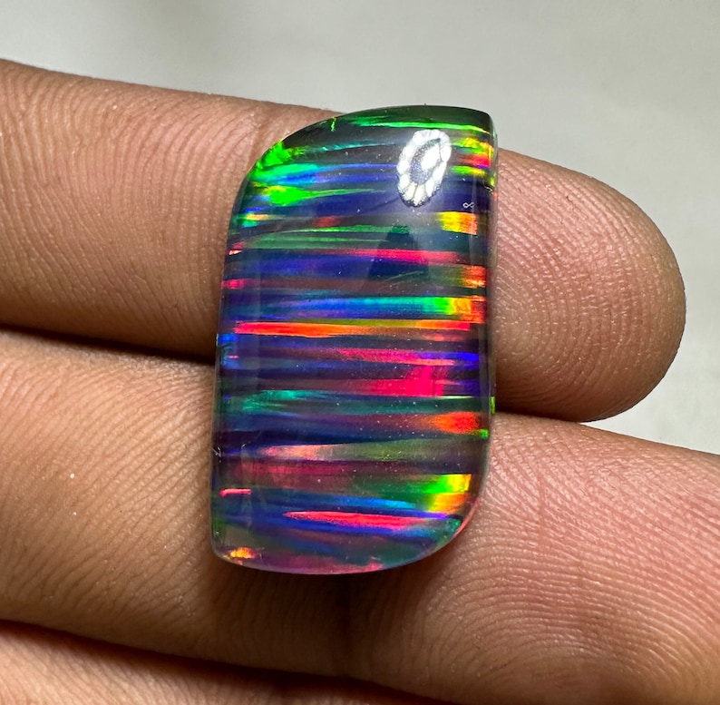 Op de afbeelding: Een zwarte opaal cabochon edelsteen met iriserende regenboogkleuren in een gestreept patroon. De steen is geslepen in een rechthoekige vorm met afgeronde hoeken.
