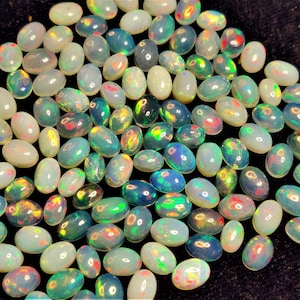 Può includere: Una collezione di opali ovali in vari colori, tra cui bianco, blu, verde e rosso. Gli opali sono sparsi su una superficie scura, mostrando il loro gioco di colori iridescenti. Queste pietre preziose sono ideali per la creazione di gioielli.