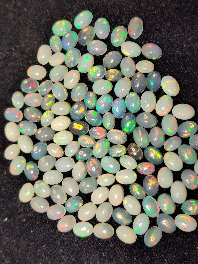 以下が含まれることがあります： 乳白色のベースカラーに、緑、黄色、赤の虹色の輝きが特徴的な、楕円形のオパールのコレクション。宝石は暗い表面に散らばっており、さまざまな色の遊びを見せています。