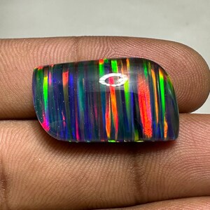 Op de afbeelding: Een cabochon geslepen zwarte opaal edelsteen met een regenboog van kleuren, waaronder rood, oranje, geel, groen, blauw en paars. De opaal heeft een gestreept patroon.