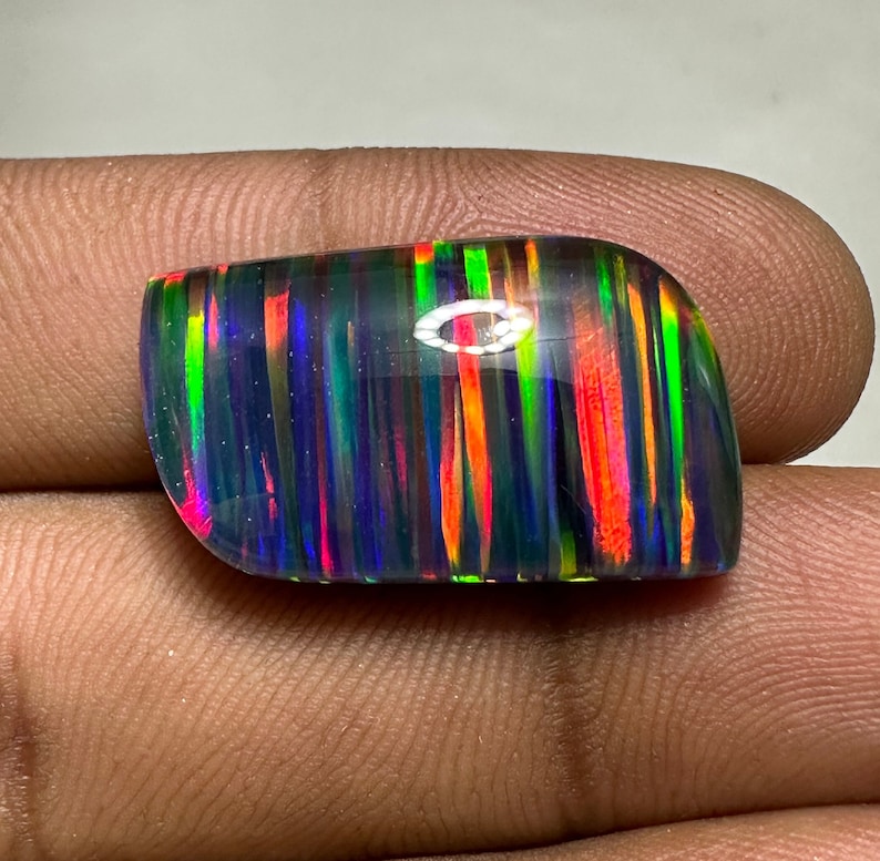 Op de afbeelding: Een cabochon geslepen zwarte opaal edelsteen met een regenboog van kleuren, waaronder rood, oranje, geel, groen, blauw en paars. De opaal heeft een gestreept patroon.