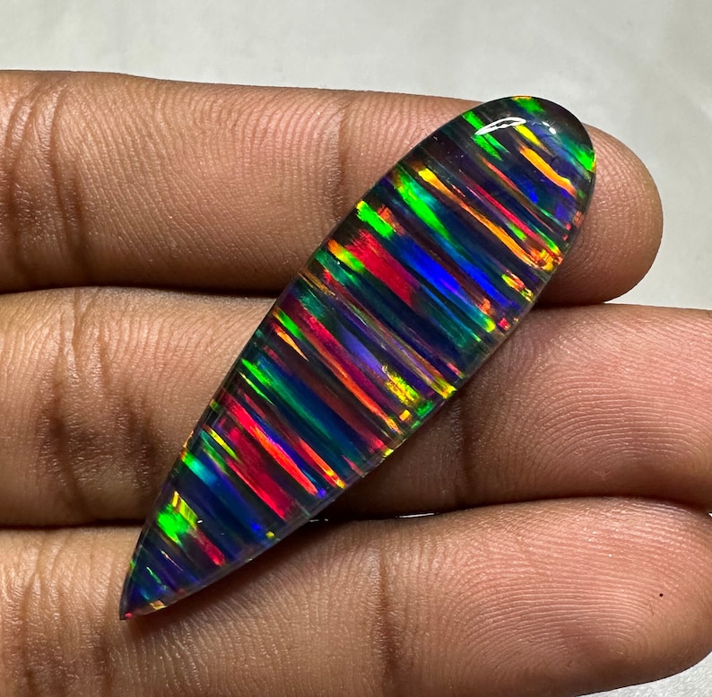 May include: A long, oval-shaped black opal gemstone with iridescent rainbow colors, including red, green, blue, and yellow.