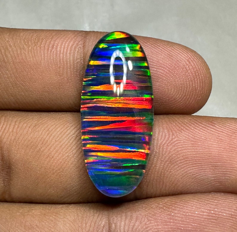 May include: Oval-shaped, iridescent opal cabochon with a rainbow of colors, including red, orange, yellow, green, blue, and purple. The opal has a black base and a striated pattern.