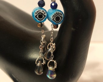 May include: Silver dangle earrings with blue and black evil eye beads, silver chain, and iridescent glass drops.