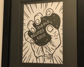 May include: Black and white illustration of a hand holding a large rock with the text "Rubble Becomes Rock" printed on it.