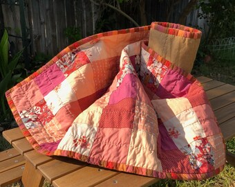 May include: A patchwork quilt with a variety of pink and orange floral patterns. The quilt is folded and placed on a wooden picnic table.