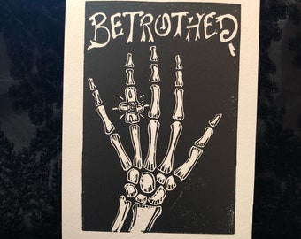 Puede incluir: Impresión en blanco y negro de una mano esquelética con un anillo, con la palabra "BETROTHED" encima. La impresión está en una tarjeta rectangular blanca. El diseño es un contraste llamativo.