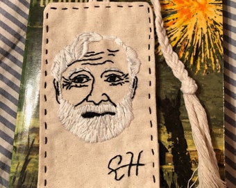 May include: A rectangular off-white fabric bookmark featuring black hand-stitched edges and a detailed white thread embroidery of a bearded man's face. The initials EH are embroidered in black at the bottom, and a braided white cord tassel is attached.