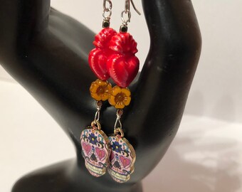 May include: A pair of dangle earrings featuring a sugar skull charm and a red heart charm. The sugar skull charm has pink and blue details, and the heart charm has a gold finish.