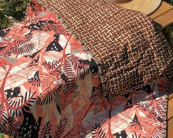 May include: A brown and pink patterned quilt with a brown and white geometric pattern on the back. The quilt is folded and placed on a wooden picnic table. A straw hat is on top of the quilt.